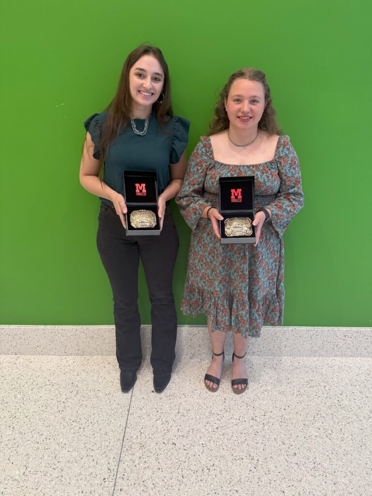 Two female students standing together in front of a green wall posing with their belt buckles